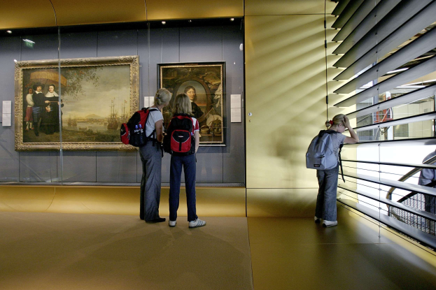 Drei Personen in einem Museum, die Gemälde an der Wand betrachten, mit einem Geländer auf der rechten Seite und Deckenlampen, die den Raum beleuchten.