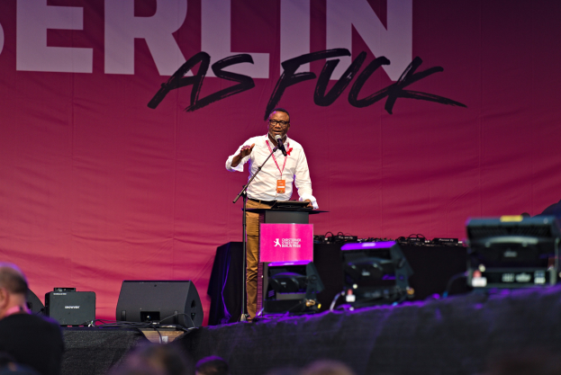 Ein Mann in einem weißen Hemd und braunen Hosen steht auf einer Bühne und hält ein Mikrofon in der Hand, mit einem pinken Banner mit der Aufschrift "Berlin as Fuck" im Hintergrund und einer Menge vor ihm.