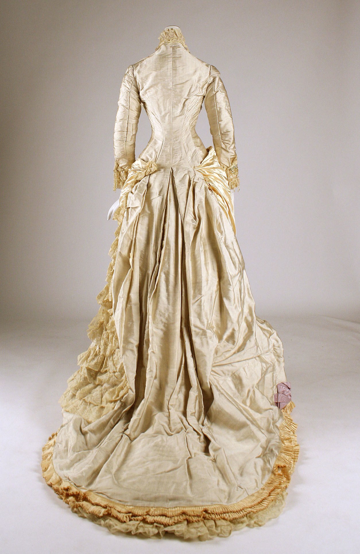 Ein cremefarbenes Ballkleid mit einer gerüschten Röcke und einem langen Zug, das auf einer Schaufensterpuppe präsentiert wird, mit einem eng anliegenden Oberteil mit Spitzenverzierung und langen Ärmeln.