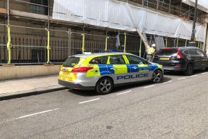 Ein Streifenwagen parkt auf einer Straße neben einem Gebäude mit zwei Helikopter-Piloten in der Nähe, einem Geländer und einem Pfahl im Vordergrund und einem weißen Tuch über dem Gebäude im Hintergrund, was auf eine Polizeiermittlung hindeutet.