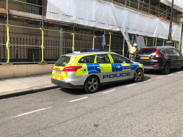 Ein Streifenwagen parkt auf einer Straße neben einem Gebäude mit zwei Helikopter-Piloten in der Nähe, einem Geländer und einem Pfahl im Vordergrund und einem weißen Tuch über dem Gebäude im Hintergrund, was auf eine Polizeiermittlung hindeutet.
