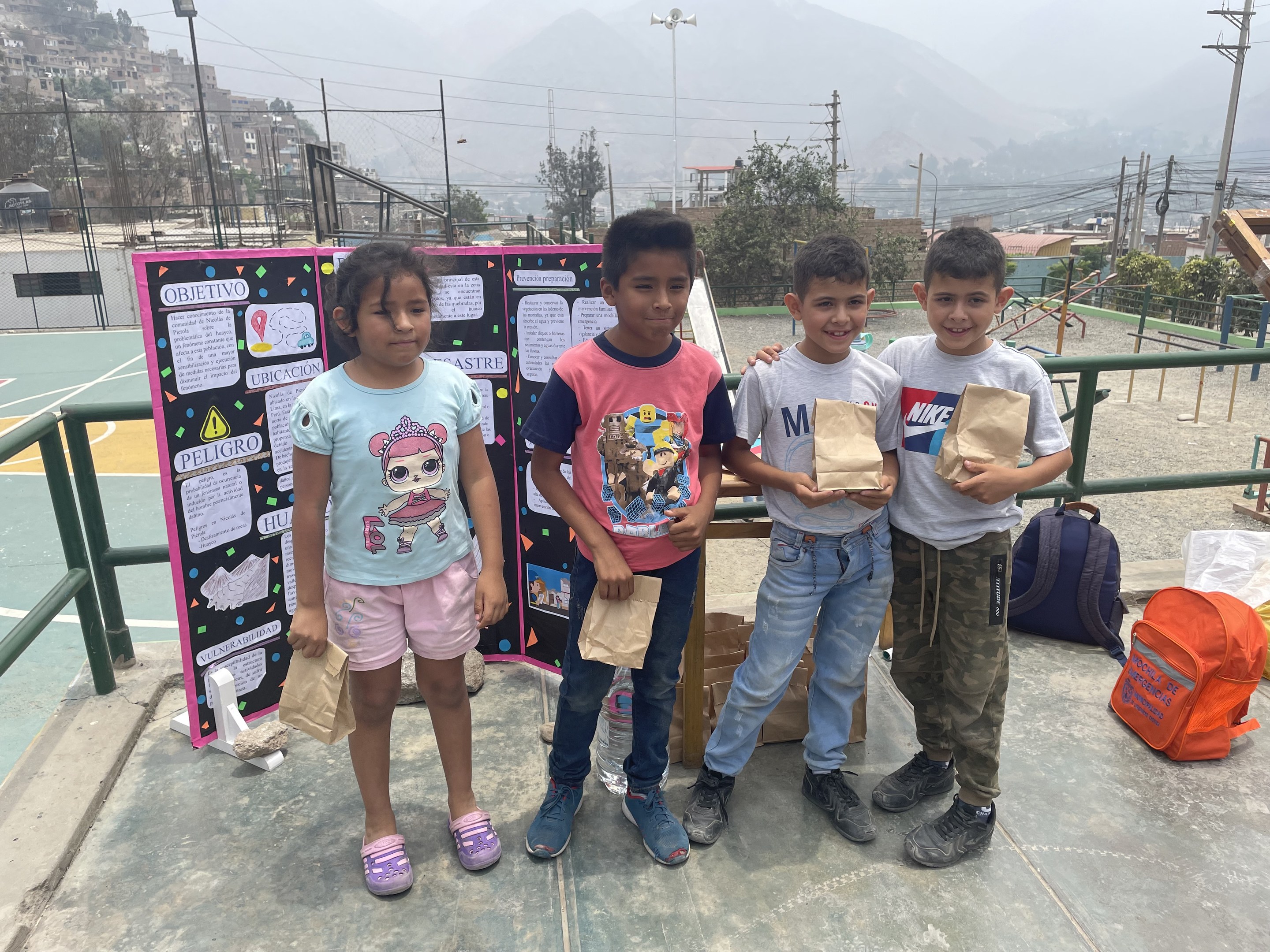 Gruppe von Kindern auf einem Basketballfeld mit Papiertüten in den Händen, mit Tüten und Gegenständen rechts und einer Tafel mit Text links, vor einer Outdoor-Background mit Zaun, Pfosten und Gebäuden.