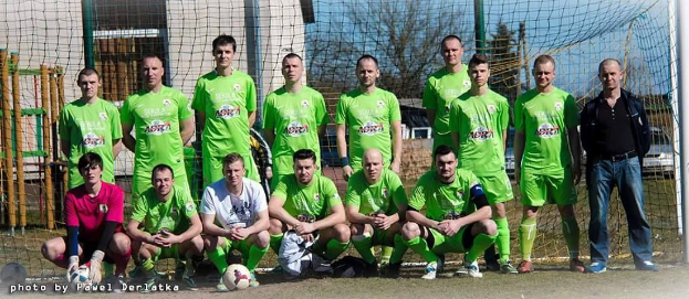 Eine Gruppe von Männern in grünen T-Shirts steht auf einem Fußballfeld in der Nähe eines Torpfostens, mit einem Fußball davor und Bäumen, Gebäuden und Fahrzeugen im Hintergrund.