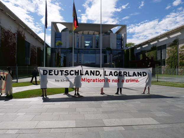 Gruppe von Menschen hält ein Transparent mit der Aufschrift "Deutschland, Lagerland Migration ist kein Verbrechen" vor einem umzäunten Gebäude mit Bäumen und Flaggen.