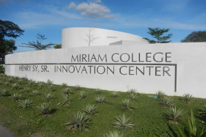 Außenansicht des Miriam College Innovation Center mit einer textbedeckten Wand, umgeben von Grün, einem Pfad, Bäumen und einem bewölkten Himmel.