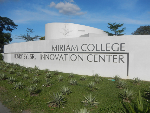 Außenansicht des Miriam College Innovation Center mit einer textbedeckten Wand, umgeben von Grün, einem Pfad, Bäumen und einem bewölkten Himmel.