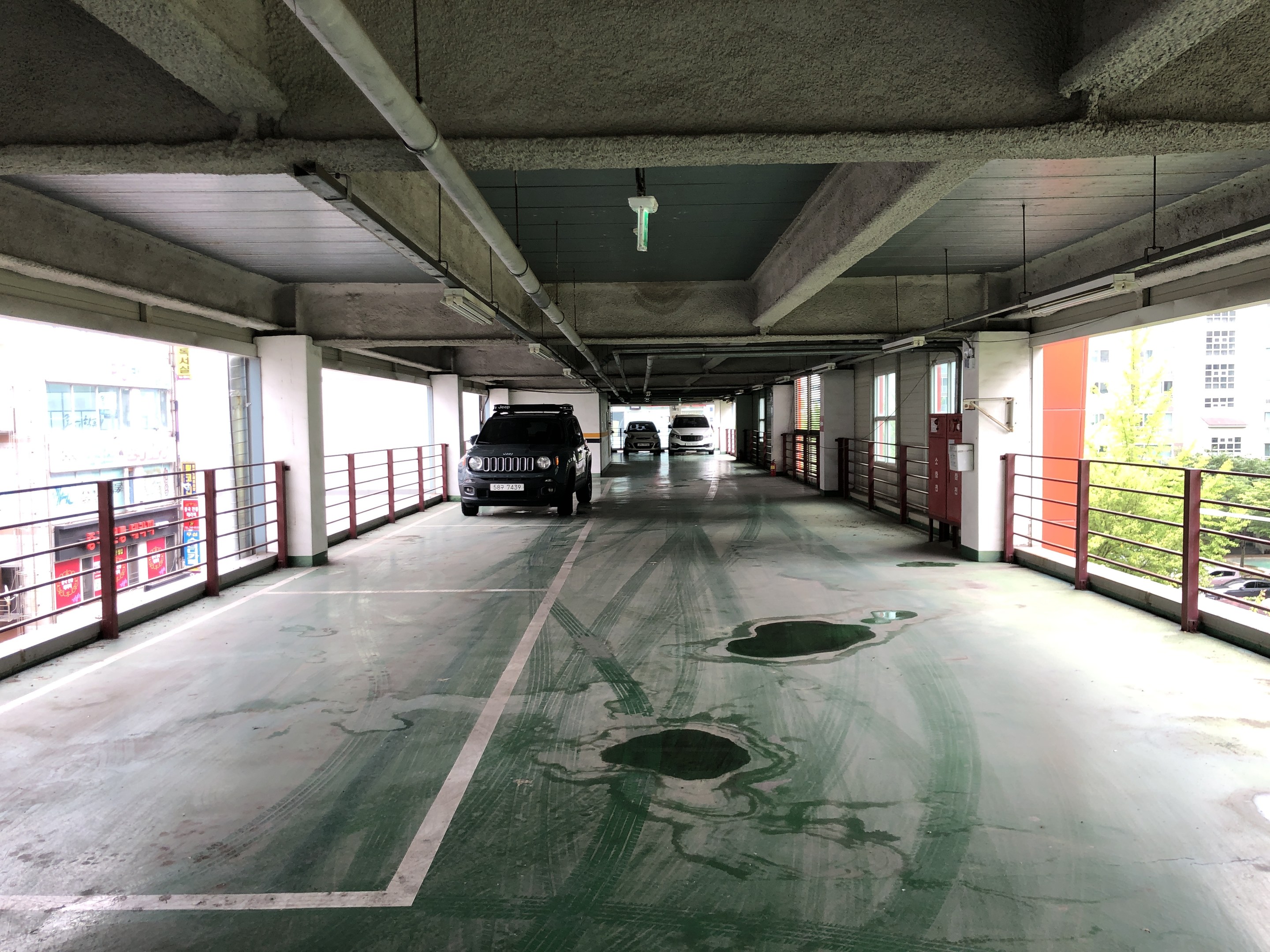 Ein mehrgeschossiges Parkhaus voller geparkter Autos, umgeben von Geländern, Säulen, Deckenleuchten und sichtbaren Rohren, mit Bäumen und einer Stadtkulisse von Gebäuden mit Fenstern im Hintergrund.