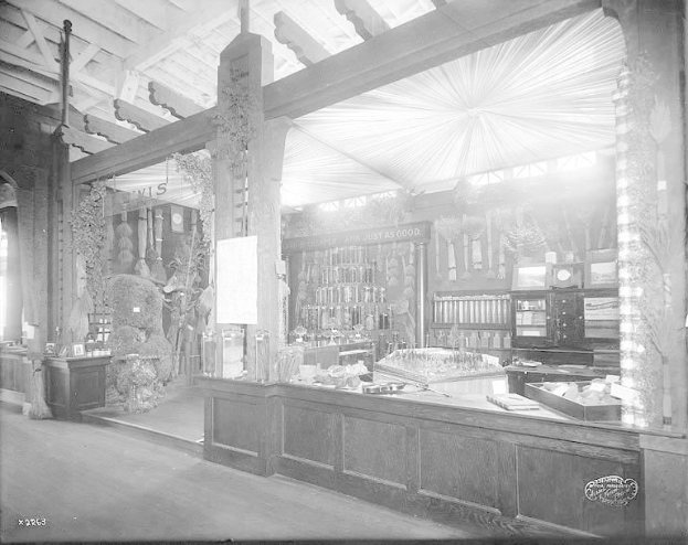 Schwarz-weißes Foto eines alten Apothekenladens mit einem zentralen Tresen, auf dem verschiedene Gegenstände ausgebreitet sind, Regalen im Hintergrund mit Flaschen und Artikeln gefüllt und Text unten, der darauf hinweist, dass es sich um eine Apotheke handelt.