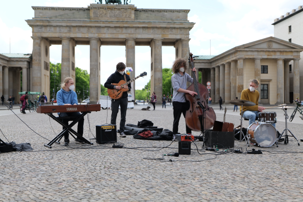 Gruppe von Menschen, die Musik im Freien spielen, mit einem Gitarristen im Sitzen, einem weiteren Musiker und einer maskierten Person; Brandenburg-Tor und städtische Elemente im Hintergrund.