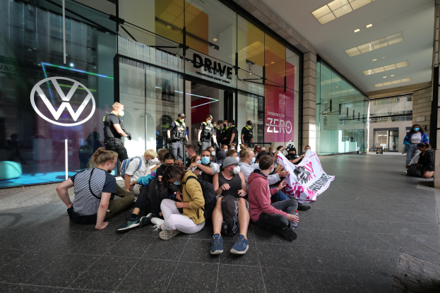 Eine Gruppe von Menschen sitzt vor einer Glasfassade eines Volkswagen-Händlers auf dem Boden und hält ein Transparent, unter Deckenleuchten, scheinbar protestierend.