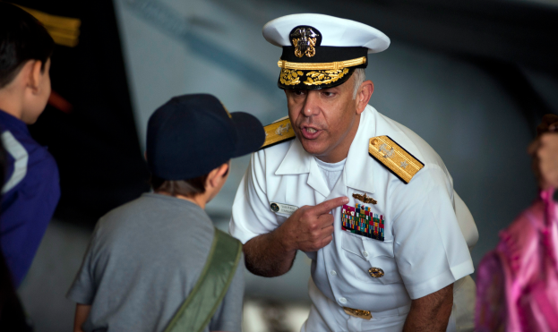 Ein Mann in wei├čer Milit├Ąruniform mit Abzeichen und M├╝tze spricht zu einer Gruppe von Kindern, mit einem unscharfen Hintergrund.