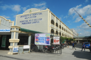 Die Universität Perpetual Help System in Pangasinan Campus mit einer Gruppe von Menschen auf dem Boden, einem Metallzaun, einer Fahne mit Text, geparkten Fahrzeugen, einem Schild, Pfosten, einem Kran, Bäumen und einem bewölkten Himmel.