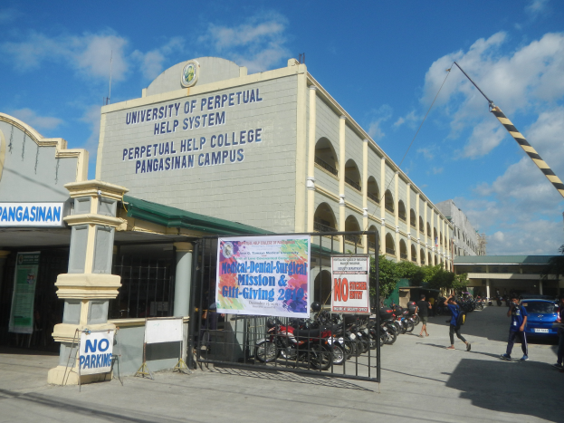 Die Universität Perpetual Help System in Pangasinan Campus mit einer Gruppe von Menschen auf dem Boden, einem Metallzaun, einer Fahne mit Text, geparkten Fahrzeugen, einem Schild, Pfosten, einem Kran, Bäumen und einem bewölkten Himmel.