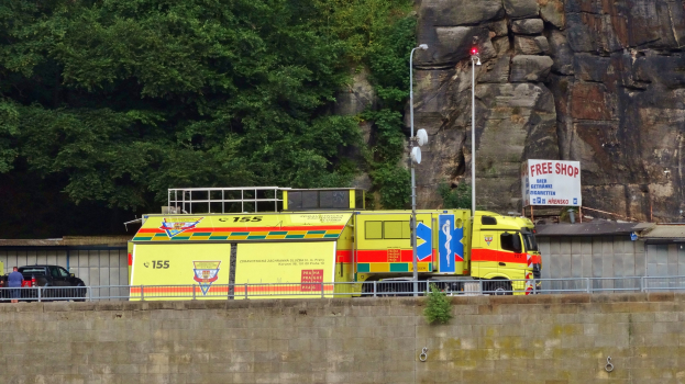 Ambulanz neben einer Straße neben einer Klippe geparkt, mit einer Wand, einem Geländer, Pfosten, Lampen und Texttafeln im Vordergrund, Bäume und ein Auto im Hintergrund.