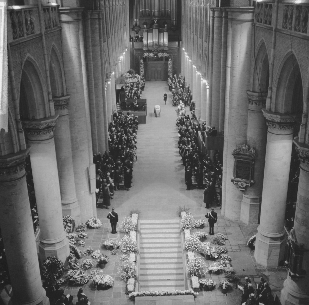 Schwarz-weißes Foto einer Trauerprozession in einer Kirche mit Menschen auf dem Boden, Blumensträuße auf beiden Seiten der Treppe zum Altar, Säulen und Lampen an den Wänden und einer Decke