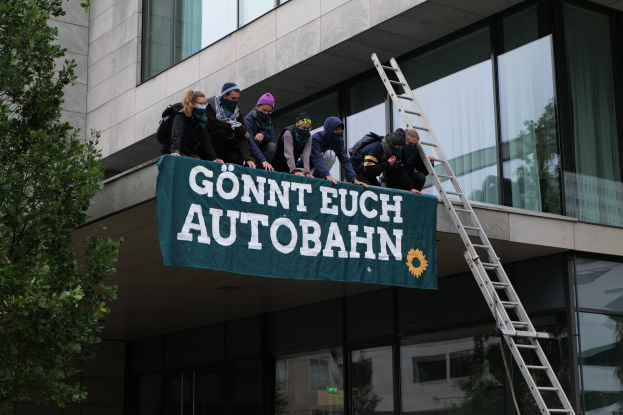 Eine Gruppe von Menschen auf dem Dach eines Gebäudes, die eine Fahne halten, mit einem Baum links und Glasfenstern im Hintergrund, die scheinbar für die deutsche Regierung protestieren.