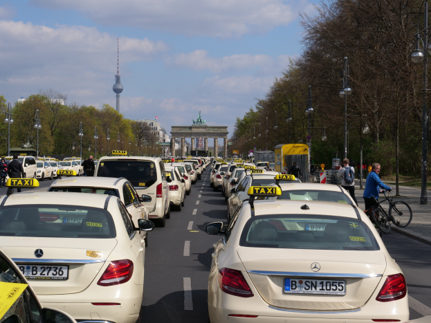 Eine lange Reihe von Taxis, die an einer belebten Straße in Berlin, Deutschland, geparkt sind, mit Fahrradfahrern und Fußgängern auf dem Gehweg, flankiert von Laternenmasten und Bäumen, und Gebäuden, einem Bogen und einem Turm im Hintergrund unter einem bewölkten Himmel.