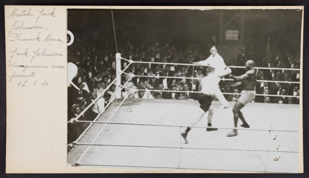 Schwarzes und weißes Foto von zwei Boxern, Jack Johnson und Frank Morgan, in einem Boxring mit Zuschauern dahinter und einer Wand im Hintergrund.
