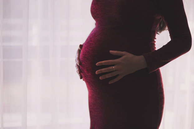Eine schwangere Frau in einem roten Kleid steht vor einem Fenster, Hände auf dem Bauch, mit leicht unscharfem Hintergrund.