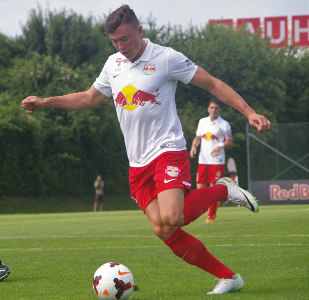 Ein Profifußballer schießt einen Ball auf einem grünen Feld mit Bäumen und einem klaren blauen Himmel im Hintergrund, in der Nähe eines Schildes mit der Aufschrift "RB Leipzig."
