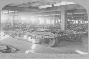 Ein Schwarz-Weiß-Foto eines Elektromotors und eines Lastwagens in einer Fabrik mit Maschinen, Säulen und einem Dach, mit Text unten.