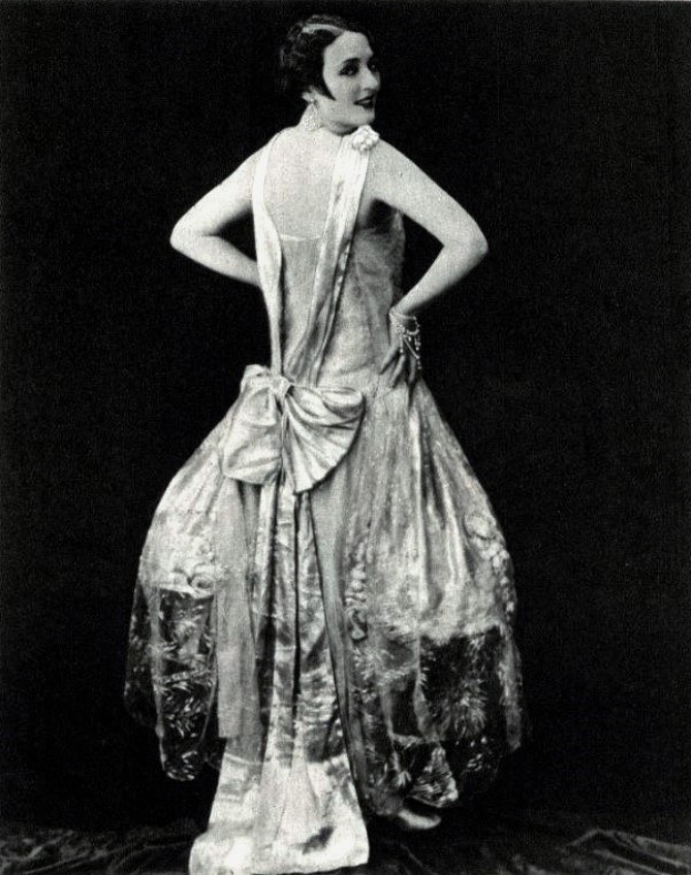 Frau in einem Kleid im Stil der 1920er Jahre mit hohem Kragen, langen Ärmeln und einem weiten Rock, die selbstbewusst mit einer eleganten Hochsteckfrisur und dezentem Make-up posiert.
