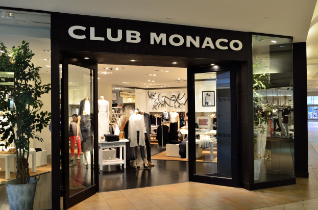 Ein Club Monaco Geschäft in einem Einkaufszentrum mit Glaswänden und -türen, das Schaufensterpuppen in Kleidern, Tische mit verschiedenen Gegenständen, einen Blumentopf und Deckenleuchten zeigt.
