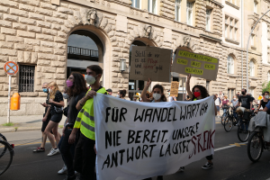 Eine Gruppe von Menschen marschiert durch eine Straße in Berlin, hält Schilder und Banner hoch und fährt teilweise Fahrräder, vor einem Gebäude mit Fenstern, Bögen, Säulen, Skulpturen, Bäumen und einem Laternenpfahl, protestiert gegen die Anti-Kriegs-Bewegung.