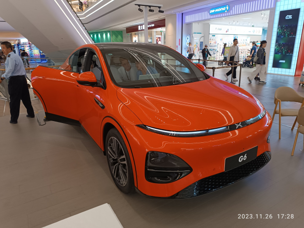 Oranges Elektroauto in einer Ausstellungshalle umgeben von Menschen, mit Stühlen und einem Wagen rechts und Geschäften im Hintergrund.