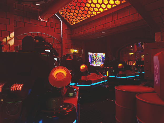 Ein hell erleuchteter Arcade-Raum voller verschiedener Videospielautomaten, Bildschirme und Gegenstände, einschließlich eines sichtbaren Rohrs und Deckenlampen.