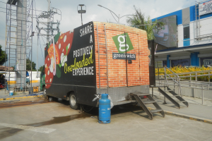 Ein Lastwagen vor einem Gebäude mit einer Tafel, auf der "Teilen Sie eine positiv überladene Erfahrung" steht, mit Stufen, einem Zylinder auf dem Boden, Geländern, Strommasten mit Drähten, Laternen, Bäumen und einem bewölkten Himmel.