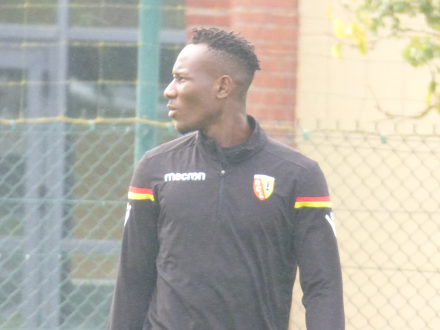 Ein Mann in einer schwarzen Jacke steht auf einem Fußballfeld, umgeben von einem Zaun und einem Baum, und sieht konzentriert während eines Trainings.