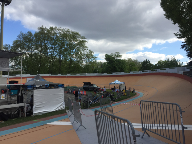 Gruppe von Radfahrern auf einer Schotterstrecke bei den UCI BMX-Weltmeisterschaften 2018, mit Zelten, Fahrzeugen, Gittern, Bäumen und einem bewölkten Himmel im Hintergrund.