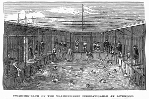 Ein Schwimmbad mit einem "Nur für Erwachsene - Nichtschwimmerbereich"-Schild im Vordergrund, umgeben von Bäumen und Gebäuden unter einem klaren blauen Himmel, mit einem Rettungsring in der Mitte des Pools.