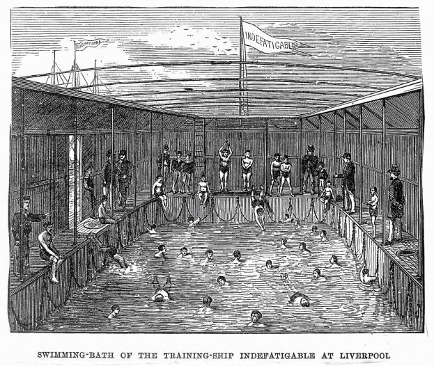 Ein Schwimmbad mit einem "Nur für Erwachsene - Nichtschwimmerbereich"-Schild im Vordergrund, umgeben von Bäumen und Gebäuden unter einem klaren blauen Himmel, mit einem Rettungsring in der Mitte des Pools.