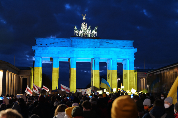 Eine Menschenmenge mit Fahnen und Plakaten steht vor dem Brandenburger Tor in Berlin, mit seinen Statuen und Säulen, die unter einem bewölkten Himmel zu sehen sind, und einem Banner mit Text auf der rechten Seite.