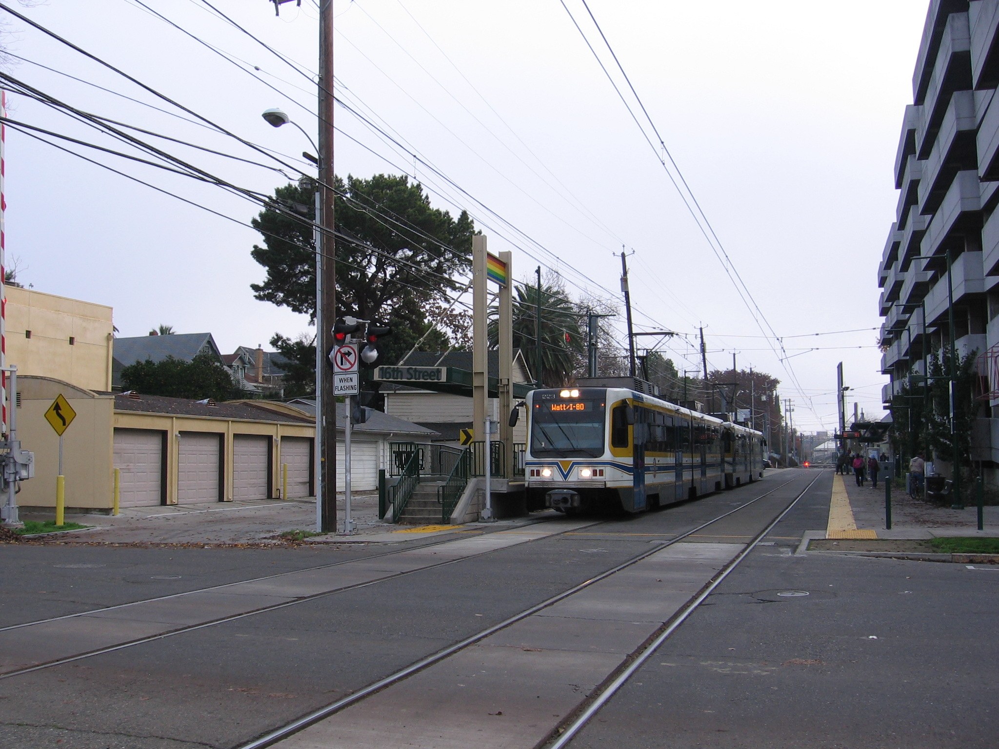 Straßenbahn fährt durch eine Straße mit hohen Gebäuden, Strommasten und Passanten, mit Bäumen und einem klaren Himmel im Hintergrund.