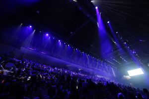 Eine große Menschenmenge vor einer Bühne mit Deckenbeleuchtung, mit einem Bildschirm auf der rechten Seite.