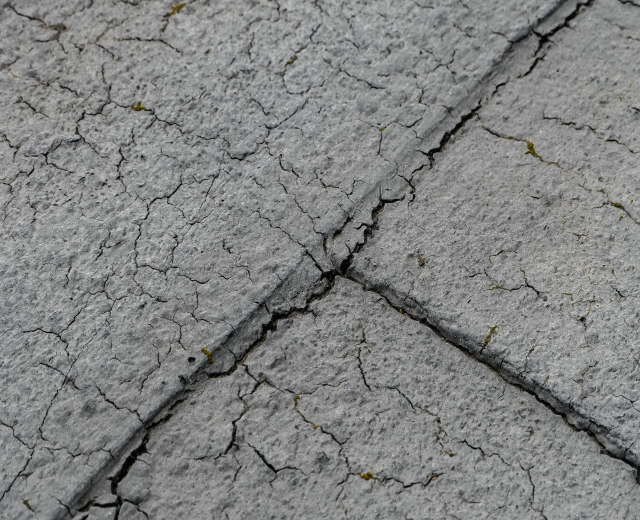 Nahaufnahme einer gebrochenen und verwitterten Straßenoberfläche mit tiefen, breiten Rissen und rauer, unebener Textur.