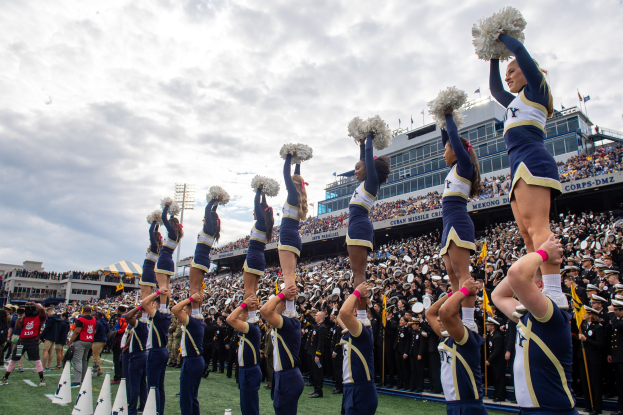 Eine Gruppe von Cheerleadern in blauen und weißen Uniformen führt einen Stunt auf einem Stadionevent durch, umgeben von Zuschauern und fotografiert von einer Person links.