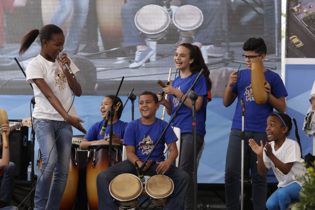 Eine Gruppe von Kindern, die auf einer Bühne Musikinstrumente spielen, mit einer Frau in der Mitte, die ein Mikrofon hält, umgeben von Mikrofonstativen und einem Banner im Hintergrund, mit Pflanzen auf der rechten Seite.