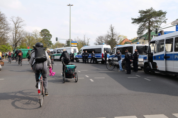 Gruppe von Menschen auf Fahrrädern auf einer Straße mit Polizeiwagen, anderen Fahrzeugen, Bäumen, Laternen, Verkehrsampeln, Verkehrszeichen, Gebäuden und einem bewölktem Himmel.