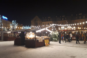 Ein belebter Weihnachtsmarkt an einem verschneiten Abend mit Menschen, Buden, Pflanzen, Gebäuden und Schildern unter einem bewölkten Himmel.