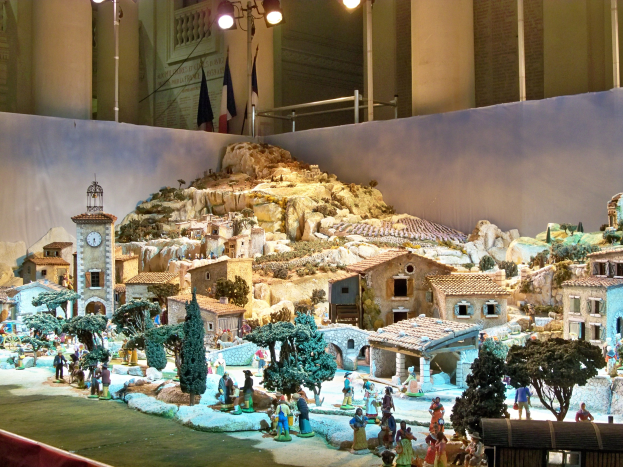 Ein detailliertes Miniatur-Modell einer Krippe in einem Museum ausgestellt, mit Figuren, Gebäuden, Bäumen, einem Kirchturm, Flaggen und beleuchtet vom Museumslicht.