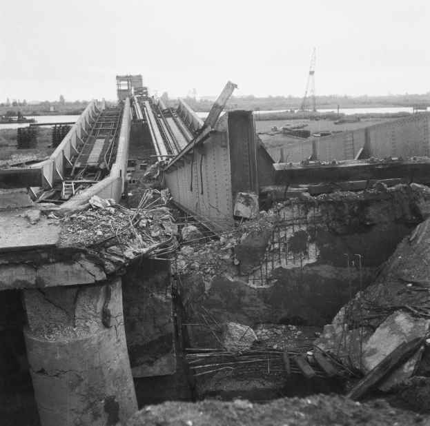 Ein Schwarz-Weiß-Foto einer teilweise gesprengten Brücke mit verstreuten Trümmern, das Wasser, Bäume und einen Turm im Hintergrund zeigt, mit dem Himmel oben.