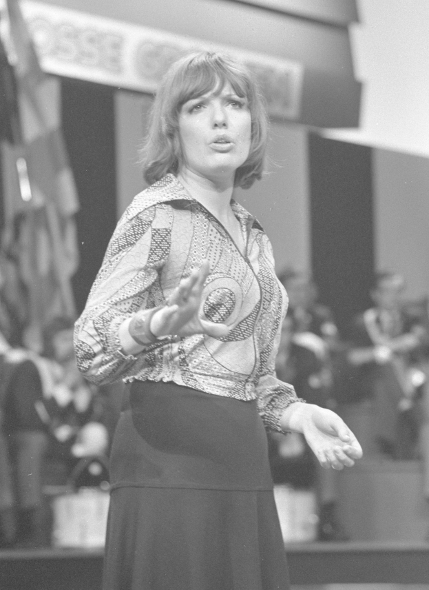 Eine Frau in einem Schwarz-Weiß-Foto steht auf einer Bühne, trägt ein weißes Hemd und einen schwarzen Rock, hat ihre Haare zu einem Dutt gebunden und einen ernsten Gesichtsausdruck, umgeben von einer Gruppe von Menschen und einer Wand im Hintergrund.