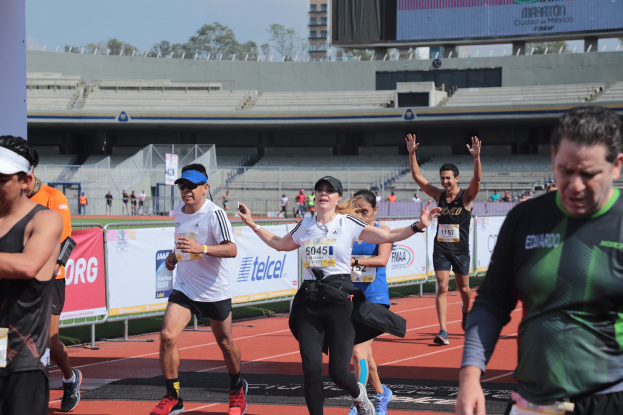 Gruppe von Läufern auf einer Outdoor-Sportanlage in einem Stadion mit Bannern, einer Anzeigetafel, einem Gebäude, Bäumen und einem bewölkten Himmel; einige Läufer tragen Turnschuhe und Hüte.