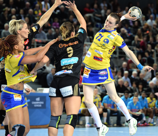 Eine Gruppe von Frauen beim Handballspielen, wobei eine den Ball hält, während unscharfe Zuschauer im Hintergrund sitzen.