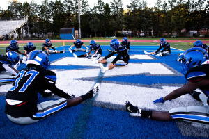 Gruppe junger Männer in Sportbekleidung, Helmen und Schuhen sitzend auf einem Fußballfeld mit Bäumen, Pfosten, Gebäuden und einem klaren blauen Himmel im Hintergrund, neben einer Treppe mit Geländern.