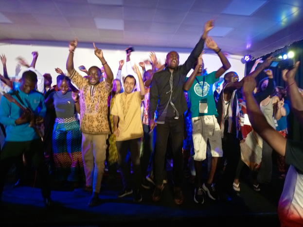 Eine Gruppe von Menschen tanzt auf einer Bühne mit erhobenen Händen, beleuchtet von Scheinwerfern auf der rechten Seite und einem Hintergrundbild.